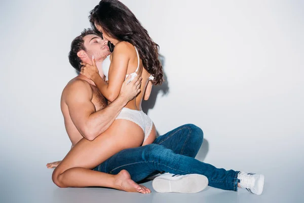 Chica seductora en lencería blanca abrazando novio sin camisa sentado sobre fondo gris - foto de stock