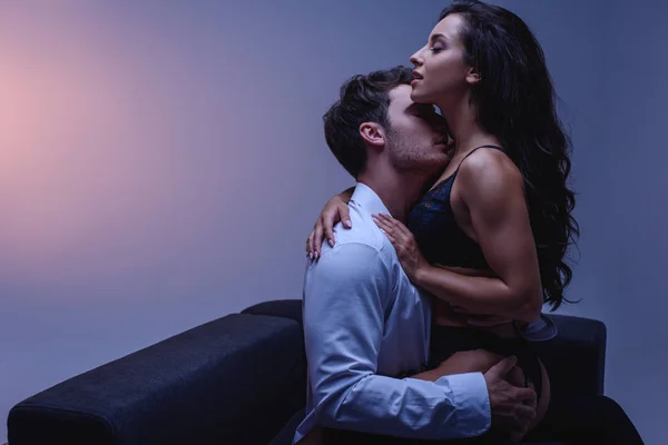Young man in white shirt hugging and kissing passionate girl in black lingerie while sitting on couch on purple background — Stock Photo