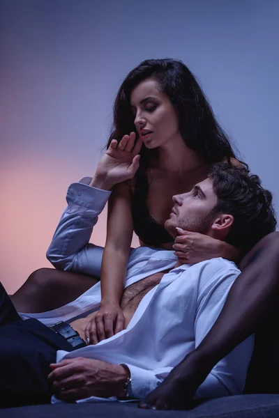 Chica seductora en sujetador negro abrazando al hombre en camisa blanca acostado en el sofá sobre fondo púrpura - foto de stock
