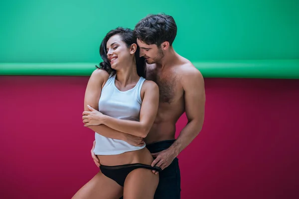 Shirtless man embracing smiling sexy girlfriend in sleeveless shirt and panties on red and green background — Stock Photo