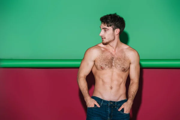 Sexy shirtless man in denim jeans holding hands in pockets and looking away on green and red background — Stock Photo