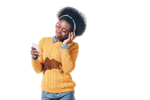 Смущенная африканская американка, использующая смартфон с наушниками, изолированная на белом — стоковое фото