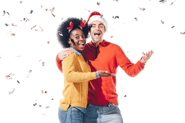 Felice coppia interrazziale in cappello di Babbo Natale e corna di cervo che celebrano con coriandoli, isolati su bianco — Foto stock