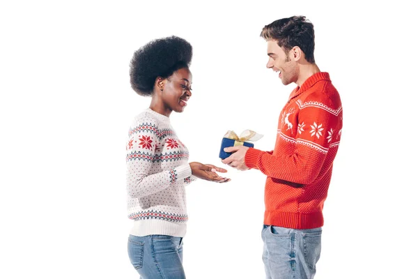 Young multiethnic couple in winter sweaters holding christmas present, isolated on white — Stock Photo
