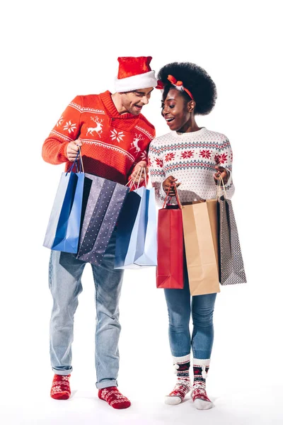 Joven pareja multicultural en sombrero de santa y cuernos de ciervo sosteniendo bolsas de compras, aislado en blanco - foto de stock