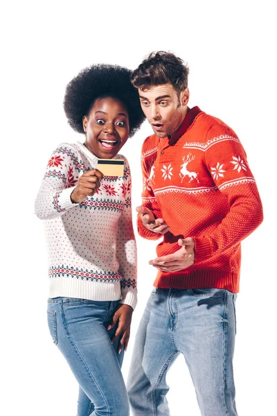 Усміхнена мультикультурна пара в зимових светрах тримає кредитну картку, ізольована на білому — стокове фото