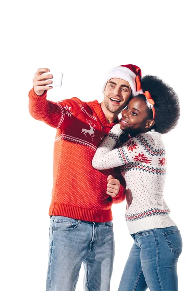 Multicultural couple in santa hat and deer horns taking selfie on smartphone, isolated on white — Stock Photo