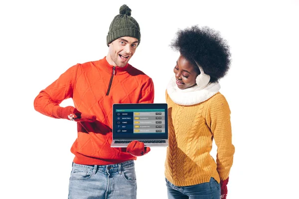 Мультикультурная пара в зимних свитерах, перчатках и шляпе держа ноутбук с погодным вебсайтом, изолированные на белом — стоковое фото
