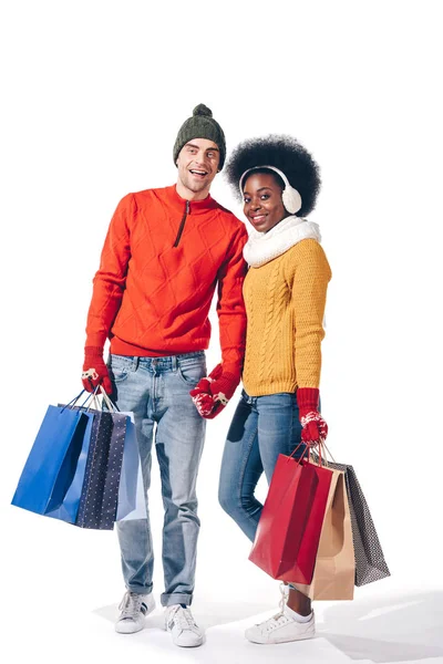 Feliz pareja multiétnica en suéteres de invierno sosteniendo bolsas de compras, aislado en blanco - foto de stock