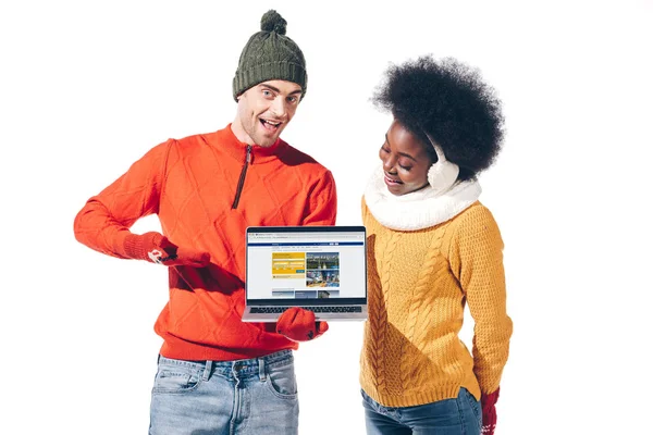 KYIV, UKRAINE - AUGUST 21, 2019: multicultural couple in winter sweaters, gloves and hat holding laptop with booking website, isolated on white — Stock Photo