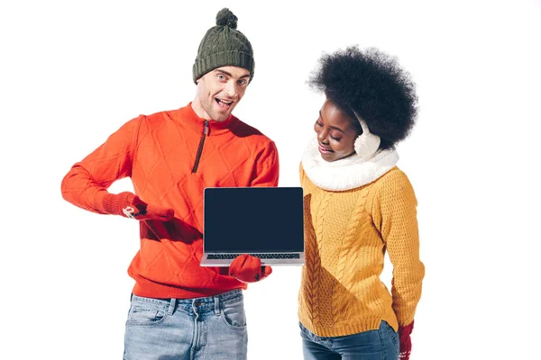 Belo casal multicultural em camisolas de inverno, luvas e chapéu segurando laptop com tela em branco, isolado em branco — Fotografia de Stock