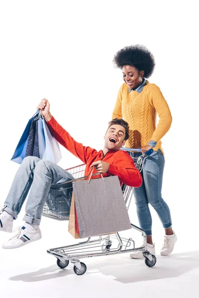 Young multicultural couple having fun in shopping cart with shopping bags, isolated on white — Stock Photo
