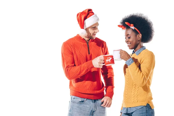 Lächelndes multikulturelles Paar mit Weihnachtsmann und Hirschhörnern in Kaffeetassen, isoliert auf weiß — Stockfoto