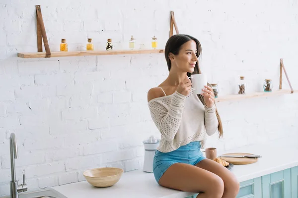Feliz joven mujer sosteniendo la taza de café en la cocina - foto de stock