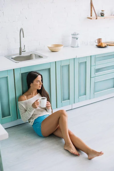 Молодая женщина с чашкой кофе сидит на полу в уютной кухне — стоковое фото