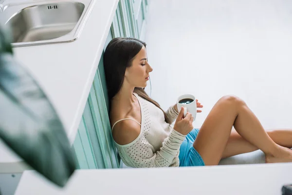 Beautiful girl sitting on floor and looking at cup of coffee in kitchen — Stock Photo