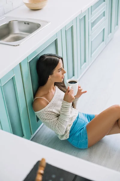 Attractive woman drinking coffee while sitting on floor in kitchen — Stock Photo