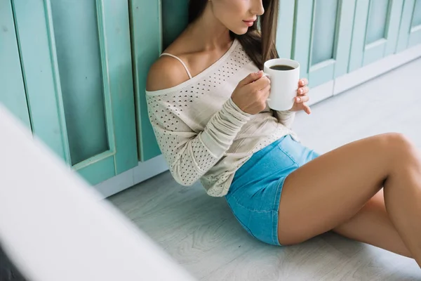 Vista recortada de la mujer sosteniendo taza de café y sentado en el suelo en la cocina - foto de stock