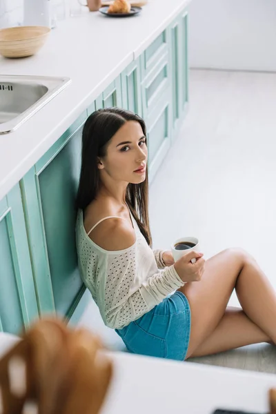 Chica atractiva con taza de café sentado en el suelo en la cocina - foto de stock