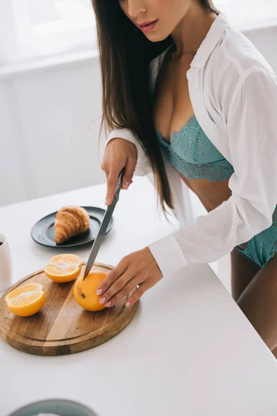 Corte vista de mulher sexy em lingerie cortando laranjas para suco na cozinha — Fotografia de Stock