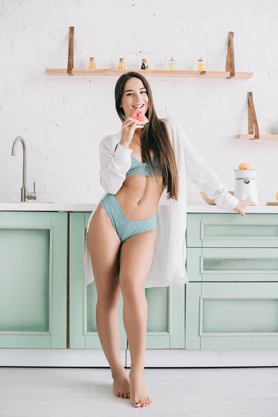 Smiling sexy girl in lingerie and white shirt holding watermelon piece in kitchen — Stock Photo