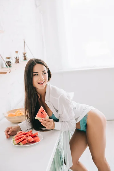 Atractiva chica feliz en lencería y camisa blanca comiendo sandía en la cocina - foto de stock