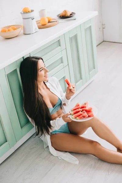 Chica sonriente en lencería y camisa blanca comiendo sandía en el suelo en la cocina - foto de stock