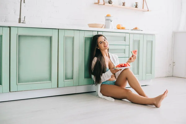 Attractive smiling girl in lingerie and white shirt eating watermelon on floor in kitchen — Stock Photo