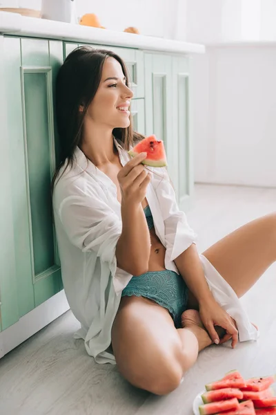 Atractiva mujer alegre en lencería y camisa blanca comiendo sandía en el suelo en la cocina - foto de stock
