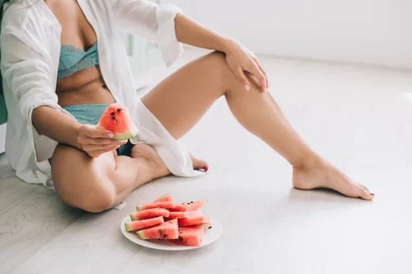 Vista ritagliata di donna in lingerie e camicia bianca mangiare pezzi di anguria sul pavimento in cucina — Foto stock