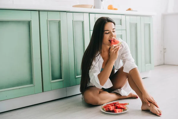 Chica sexy en lencería y camisa blanca comiendo piezas de sandía en el suelo en la cocina - foto de stock