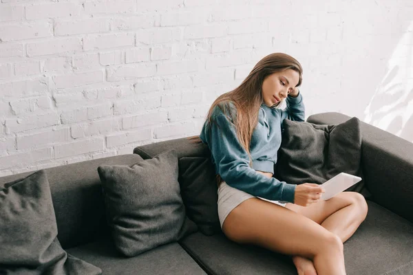 Atractiva chica pensativa en bragas usando tableta digital en el sofá - foto de stock