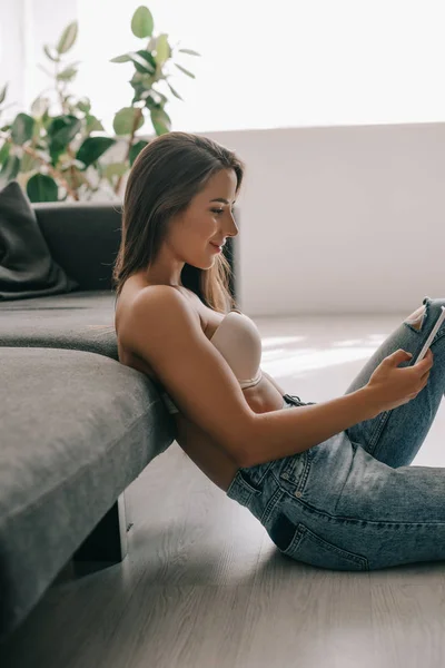 Приваблива усміхнена дівчина в білому бюстгальтері і джинсах, використовуючи смартфон, сидячи на підлозі біля дивана — стокове фото