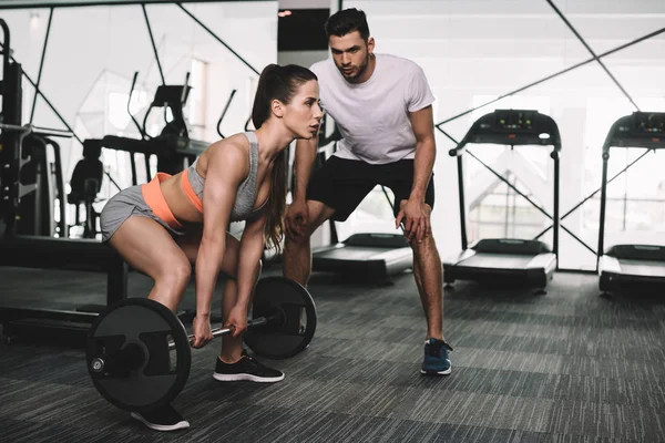 Handsome trainer looking at athletic sportswoman lifting barbell — Stock Photo