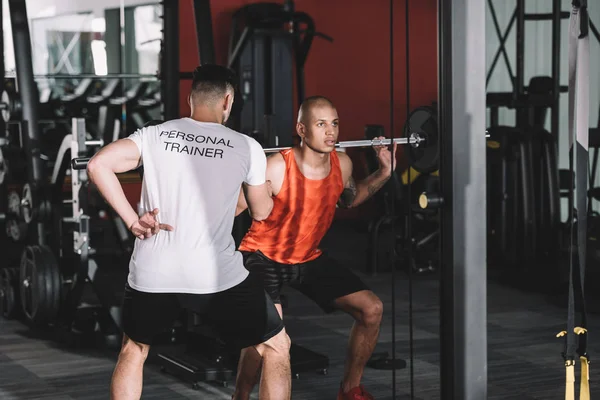 Visão traseira do personal trainer controlando jovem esportista afro-americano levantando barbell — Fotografia de Stock