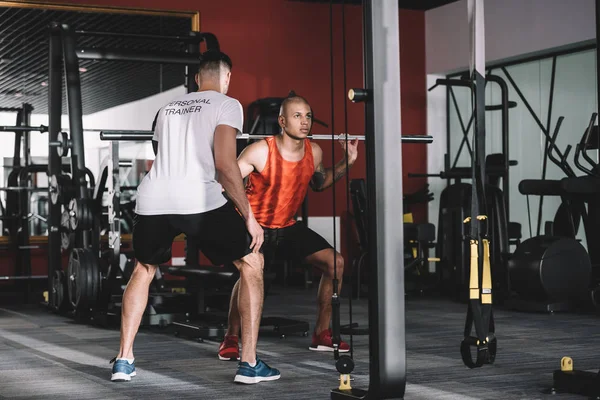 Visão traseira do personal trainer supervisionando jovem esportista afro-americano levantando barbell — Fotografia de Stock