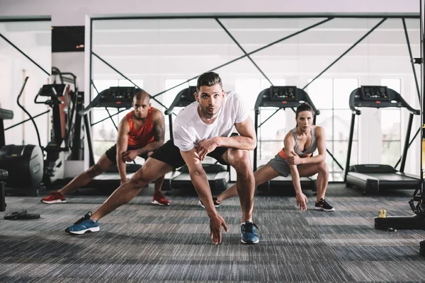 Schöner Trainer, der in die Kamera schaut, während er mit multikulturellen Athleten Ausfallübungen macht — Stockfoto