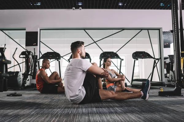 Young trainer with multicultural athletes stretching while sitting on floor in gym — Stock Photo