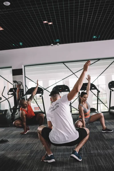 Back view of trainer squatting with raised hand together with multicultural athletes — Stock Photo