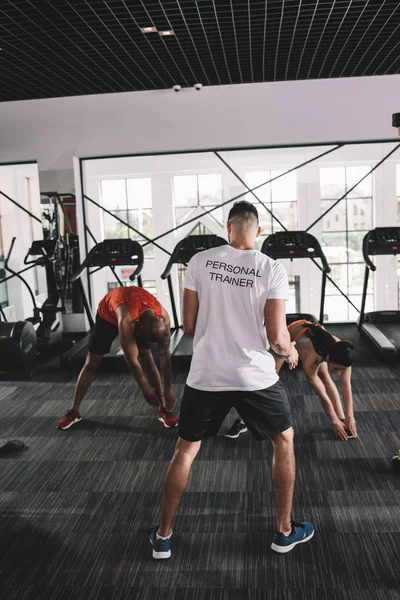 Vue de dos de l'entraîneur personnel supervisant des athlètes multiculturels s'étirant dans la salle de gym — Photo de stock