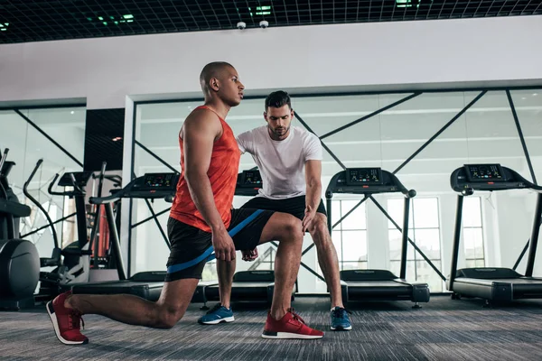 Attentive trainer controlling african american sportsman exercising with resistance band — Stock Photo