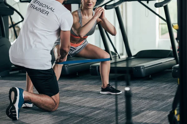Vista recortada del entrenador personal instruyendo a la joven deportista haciendo ejercicio con banda de resistencia - foto de stock