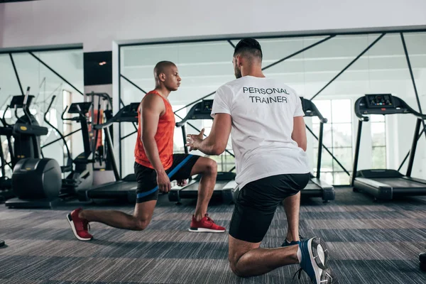 Vista posterior del entrenador personal que instruye al deportista afroamericano que hace ejercicio con banda de resistencia - foto de stock