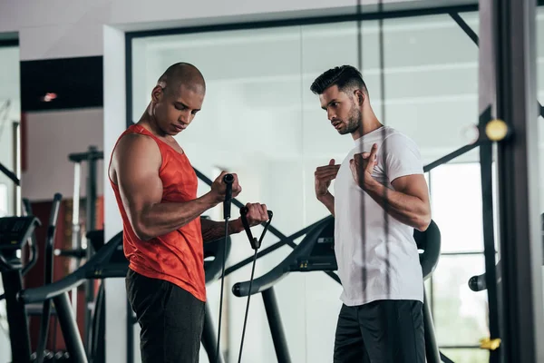 Young trainer instructing african american sportsman exercising with expander — Stock Photo