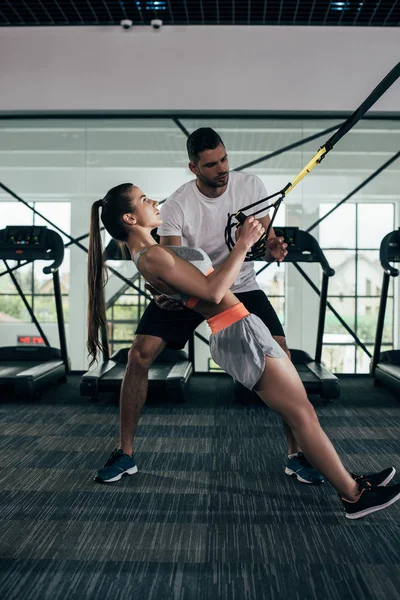 Attentive trainer supporting young sportswoman working out on suspension trainer — Stock Photo