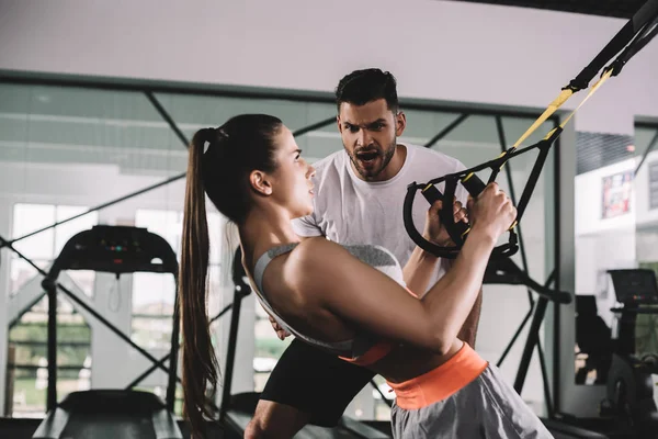 Cropped view of trainer supporting young sportswoman pulling up on suspension trainer — Stock Photo