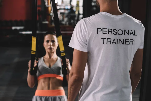 Cropped view of personal trainer standing near young sportswoman pulling up on suspension trainer — Stock Photo