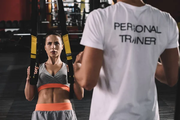 Partial view of personal trainer standing near young sportswoman pulling up on suspension trainer — Stock Photo