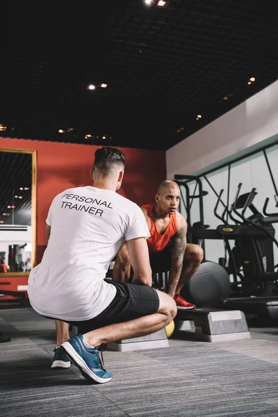 Visão traseira do personal trainer supervisionando o peso de levantamento de atleta afro-americano no ginásio — Fotografia de Stock