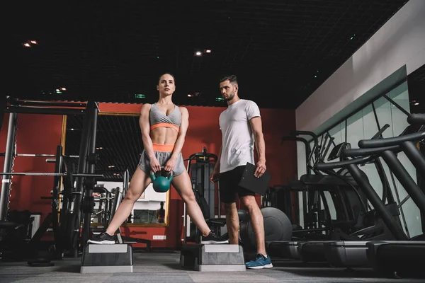 Aufmerksame Trainerin beobachtet junge Sportlerin beim Gewichtheben im Fitnessstudio — Stockfoto